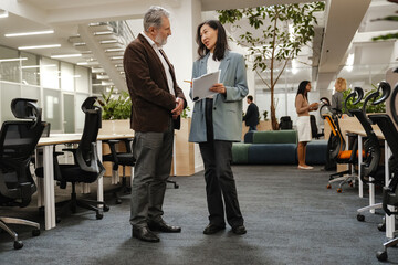 A White elder male office worker and his Asian female colleague are discussing ideas, strategies and reviewing documents in a modern office space, also with colleagues working in the background.