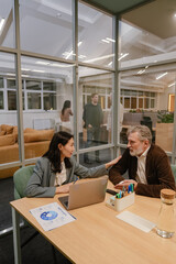 A senior man in a brown blazer and an Asian woman in a blue blazer are having a discussion in a modern office meeting room. She places a hand on his shoulder while they talk at a wooden desk.