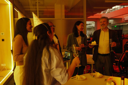 A diverse group of adults, including an elderly White man, a middle-aged Asian woman, and young Black and Hispanic women, celebrating in an office with cake and champagne. The group is chatting.