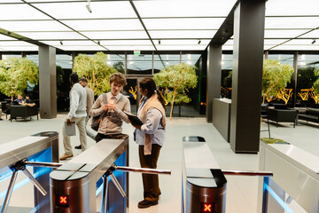A White young man and an Indian young woman discussing details during an internship meeting in a modern office lobby, with turnstiles and lush plants in the background, engaged in conversation.