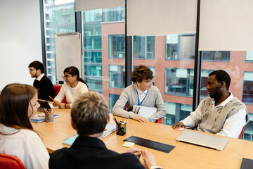 A diverse group of young professionals sits around a conference table in a modern office, engaged in a discussion. A young White man in a lanyard talks with a young Black man.