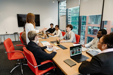 A diverse group of young professionals gathered around a conference table in a modern office. A young White woman stands leading the discussion, while colleagues engage in the meeting.