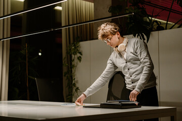 A young White man with glasses and headphones around his neck organizes sticky notes at a sleek white desk in a modern office at night. He wears a light gray cardigan and is focused on his task.
