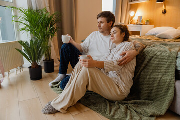 A White man and a pregnant woman in pajamas are sitting on the floor at home, embracing and drinking coffee in the morning. The couple is enjoying a peaceful moment together in a cozy bedroom setting.