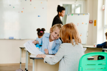 Two school-aged boys sit at the desks in a classroom, smiling and sharing drawings. A Black female teacher stands in the background near a whiteboard, while other students focus on their activities.