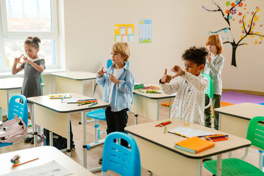 Four children, including a Black girl, Caucasian boy, multiracial boy, and red-haired Caucasian girl, learning sign language in a classroom. They are standing near desks with school supplies.