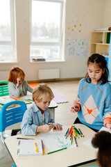 White boy in a light blue shirt focuses on drawing with colored pencils, while a girl in a blue sweater stands nearby holding a red pencil. Other students sit at desks, engaged in creative activities.