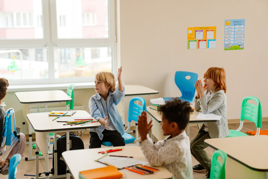 boy in a light blue shirt eagerly raises his hand while participating in a classroom discussion. Other students, seated at modern desks with colorful school supplies, engage in learning activities.