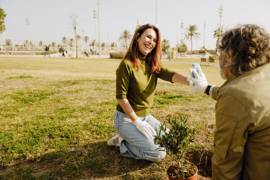 A White adult woman smiling and kneeling in a park while planting an olive tree, giving a high-five to a White elder man, as they enjoy a sunny day together outdoors. - Powered by Adobe