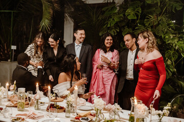 A Black groom and bride celebrating with a multinational group of young people, enjoying drinks and dancing at a wedding reception surrounded by candles, flowers, and food in an elegant venue.