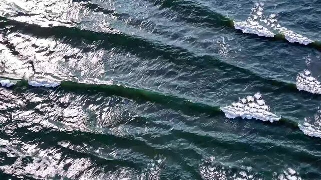 Aerial view of ocean waves breaking in perfect parallel lines, creating a mesmerizing natural pattern under the sunlight.