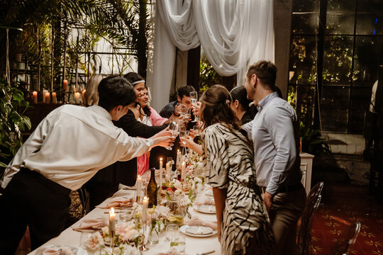 A multinational group of young people are celebrating and toasting with champagne glasses at a wedding reception, surrounded by candles, flowers, and a beautifully decorated table in an elegant venue.