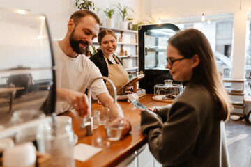 A friendly barista and a woman customer interacting at the counter of a cozy cafe, with the barista serving water while another barista smiles in the background, creating a welcoming atmosphere.