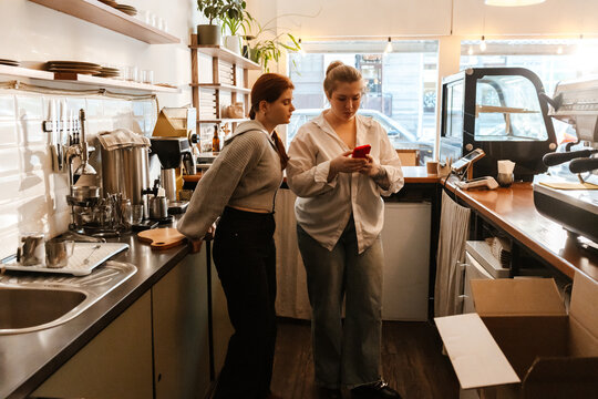 Two adult women stand behind the counter of a modern cafe, engaged in a discussion while looking at a smartphone. One woman wears a white blouse and jeans, while the other leans on the counter.