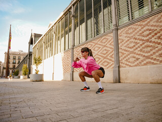 A White young woman performing squats outdoors on a sunny day, wearing a pink sweatshirt, shorts, and sneakers, engaged in a fitness workout in an urban environment with a brick wall backdrop.