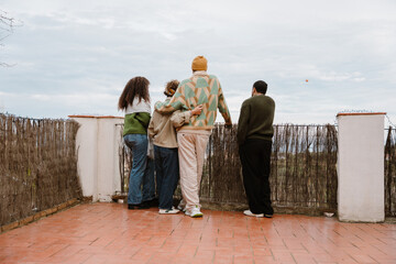 A group of young people stands on a terrace, gazing at the distant landscape. A man in a beanie...