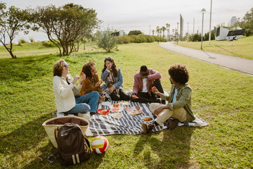 Group of five adults sitting on a picnic blanket in a park during daytime, eating fruit and snacks. A Black man with an afro wears a green jacket, a white man with curly hair wears a cream jacket.