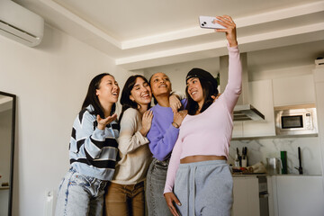 A multinational group of young women posing for a selfie together in a modern kitchen, laughing and...