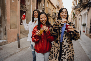 Three young women stroll down a street, laughing and enjoying colorful drinks. Their stylish...