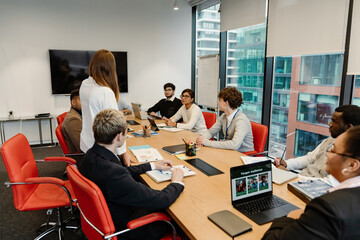 A female employee stands at a table with a group of six employees and a male intern sitting at it, in an office, during an internship