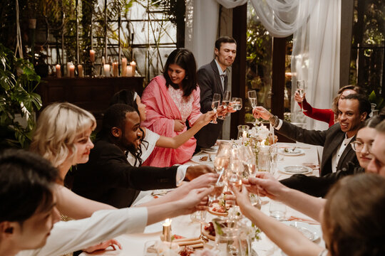 Bridesmaids stand and clink glasses with guests and the bride and groom sitting next to them at a table, in an establishment, at a wedding celebration