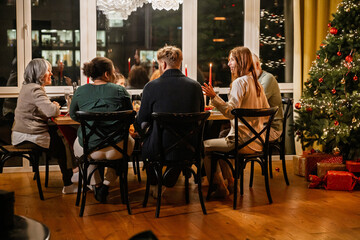 Family and guests sitting at the table and talking while looking at each other, in the living room, during Christmas dinner