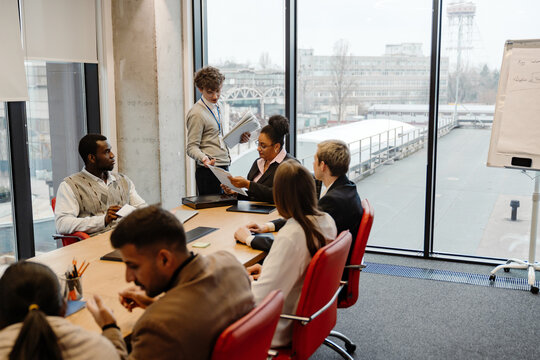 A male intern walks and hands out clipboards to a group of six employees sitting next to him at a table in an office during an internship