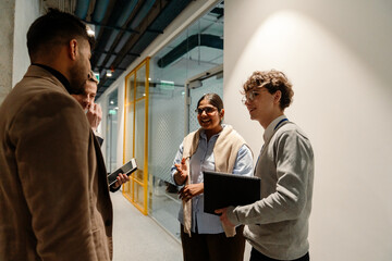 Group of three employees and a male intern stand and smile while talking and holding a notebook and...