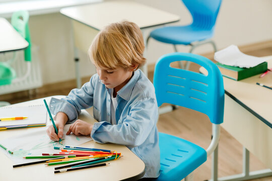 A schoolboy sits at a desk and draws with a pencil, at school, in the classroom - Powered by Adobe