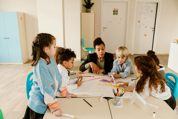 A group of six schoolchildren stand and sit at desks while drawing and listening to a teacher...