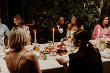 A group of five guests and the bride and groom are sitting at a table and talking, during a meal, in a restaurant, at a wedding