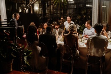 A male guest stands and smiles while holding a glass and looking towards the bride and groom and a...