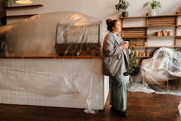 Female owner standing and leaning on the bar while holding a cup and looking ahead, preparing to...