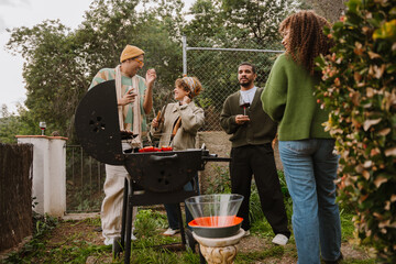 Group of four friends standing and laughing while holding glasses and tongs, outdoors near a grill, during dinner
