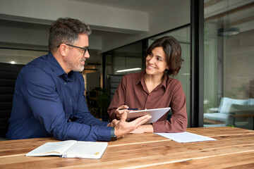 Professional business people consulting and collaborating sitting in office using digital tablet. Two happy busy mid age business man and young woman executives partners team working together on tab.