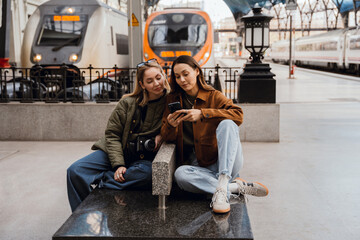 A woman sits and looks at the phone held by her female friend sitting next to her, at a train station, while traveling