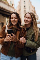 A woman stands and looks at the phone held by her friend standing next to her, they are laughing, on the street, while traveling