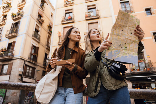 A woman stands and smiles while holding street food, with a female friend standing next to her holding a map, on the street, while traveling