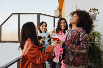 Group of four female friends standing and laughing while holding glasses, on a balcony, at a girls'...