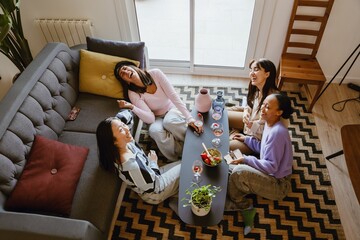 Top view of a group of four female friends sitting at a table and laughing while drinking, in the living room, at a girls' party