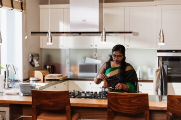 A woman stands and holds a wooden spoon over a pan on the stove while preparing dinner, in the...