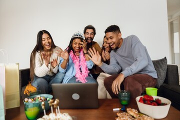 A group of five friends are sitting on a sofa and laughing while looking at and waving at a laptop in front of them on a table, at a birthday party