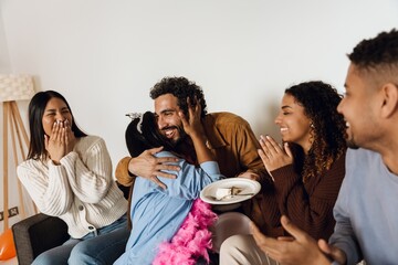 A woman sits and hugs a man sitting next to her and holds a plate while a group of three friends sit next to them and applaud, at a birthday party