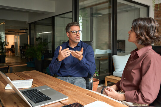 Male executive manager explaining corporate project plan to female partner colleague sitting at office desk. Business team of two busy professionals working together discussing corporate strategy.