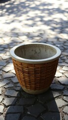 Naklejka premium Wicker-textured concrete pot sits on cracked asphalt, dappled sunlight, asphalt, detail, empty