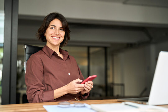 Young happy business woman holding smartphone using cellphone at work looking away. Smiling businesswoman checking mobile apps ai solutions on phone while working sitting at office desk.