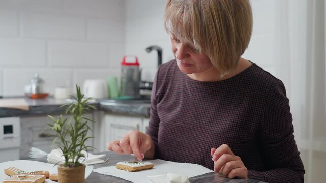 Close-up of woman placing green decorative bead on cookie while decorating, with floral arrangement, baking essentials, and kitchen equipment in background