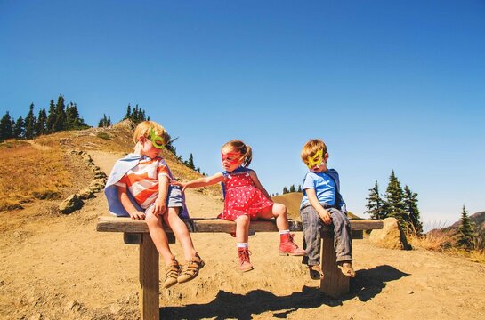 Three children dressed as superheroes sitting on a bench on a summer day