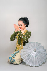 Indonesian woman in a traditional kebaya and batik, gracefully kneeling with a vintage parasol. She...