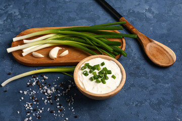 Bowl of tasty sour cream and board with green onion on blue background